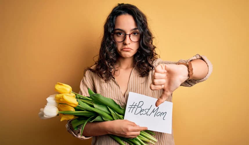 Gözlüklü genç anne, elinde sarı laleler ve “#BestMom” yazılı kâğıtla üzgün bir ifadeyle başparmağını aşağı gösteriyor; mükemmel anne baskısı ve yetersizlik hissini simgeleyen görsel