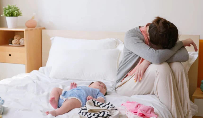 Yatakta mavi tulumlu bebek uyurken, yatağın kenarında başını kollarına dayamış yorgun görünen anne; etrafta bebek kıyafetleri ve ev ortamı