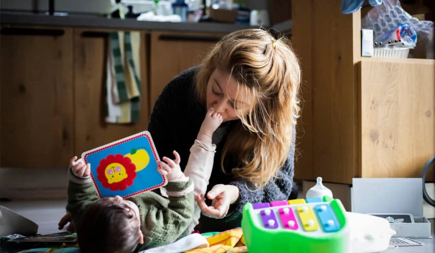 Evin mutfak kısmında yerde oynayan bir anne ve bebek; anne aslan resimli renkli bir aktivite kitabı tutarken bebek yüzüne dokunuyor, etrafta oyuncaklar ve biberon var